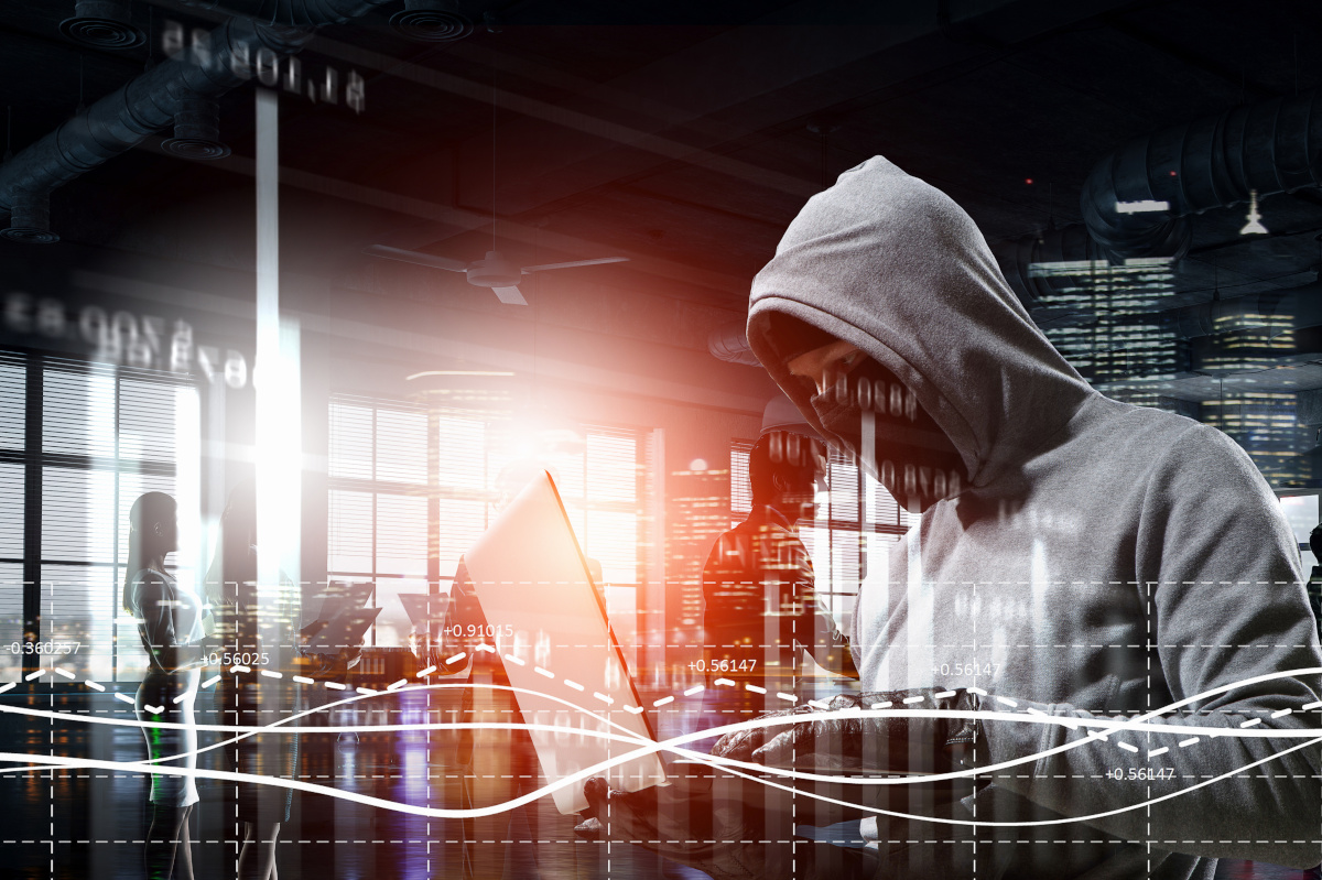 A man in a gray hoodie wearing a mask and gloves typing on a laptop with data overlaid on top the image to represent hacking